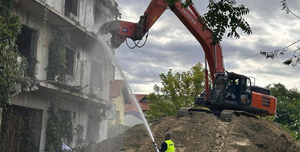 Hitachi lánctalpas kotró törőollóval bont egy elhagyott, többszintes vasbeton épületet, előtérben vízsugaras porkötés